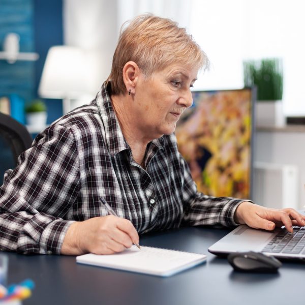 Mature entrepreneur taking notes on notebook working in home office. Elderly woman in home living room using moder technoloy laptop for communication sitting at desk indoors.